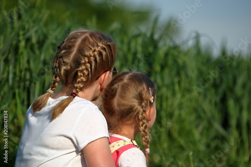 Dwie dziewczynki, blondynki uczesane w warkoczyki, nastolatka i kilkulatka, siedzą razem, blisko siebie, na zewnątrz, tyłem, w tle  bujna wiosenna (letnia) zieleń