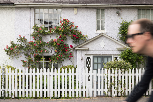 Stone white cottage with a front garden and beautiful blooming rose. A man on a bicycle is moving along a road in the background of house - out of focus. English lifestyle. Concept: travel. 
