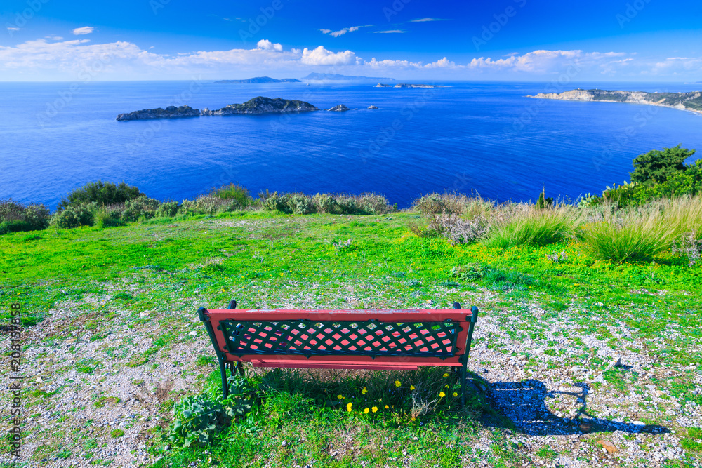 Awesome view from red wooden bench to the coastline sea bay with crystal clear azure water, distant islands in the calm warm sea. Beautiful summer seascape. Agios Stefanos cape. Afionas. Corfu. Greece