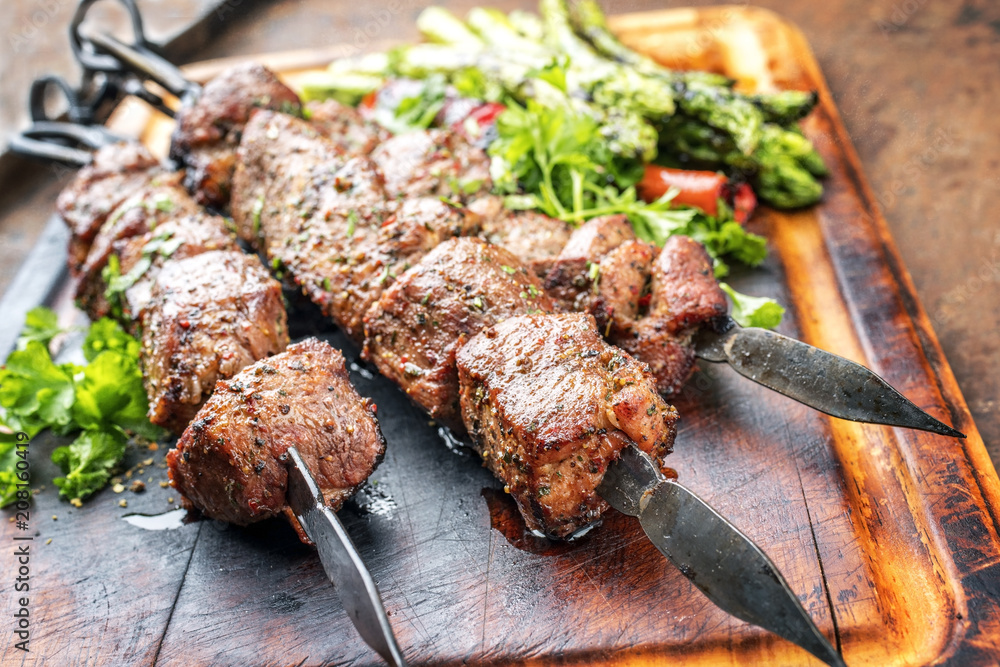 Traditional Russian shashlik on a barbecue skewer with green asparagus and paprika as close up on a burnt cutting board