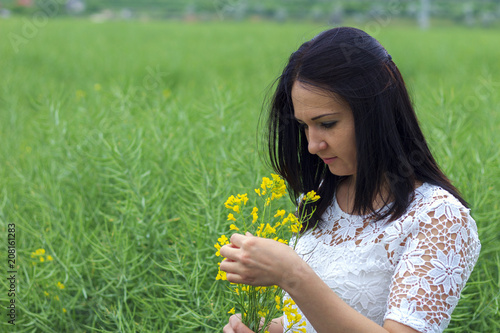 A beautiful young brunette is looking at a bouquet of yellow rape flowers in a green field . photo