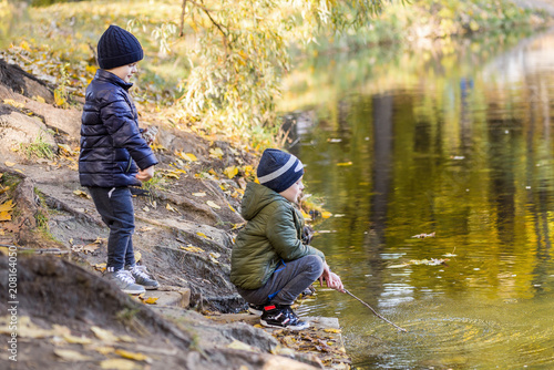 Two young boys playing fishing with sticks near pond in fall park. Little brothers having fun near lake or river in autumn. Happy childhood and children outdoor recreation concept