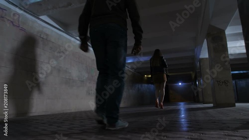 Close-up male attacker legs following young female in mini skirt at night in dark pedestrian underpass. Scared female looking back at criminal and starts running. Steadicam shot