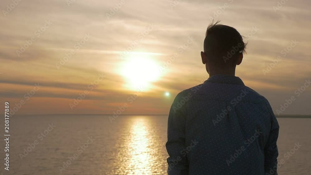 Back view of an emotional young man in sunglasses standing and staring at a picturesque sun path on the Black Sea shore at a splendid sunset in summer Back view of an emotional young man in sunglasses standing and staring at a picturesque sun path on the Black Sea shore at a splendid sunset in summer