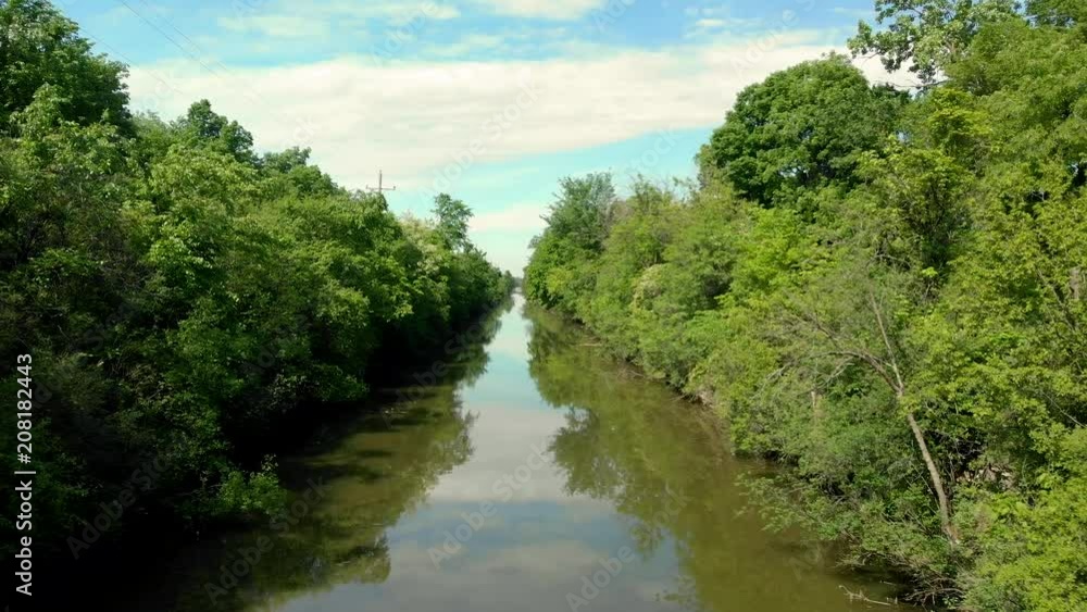 drone flying down the middle of the historic Illinois and Michigan canal with bright green trees on the side and bright blue sky with clouds in the sky and murky brown water underneath. drone flying down the middle of the historic Illinois and Michigan canal with bright green trees on the side and bright blue sky with clouds in the sky and murky brown water underneath.
