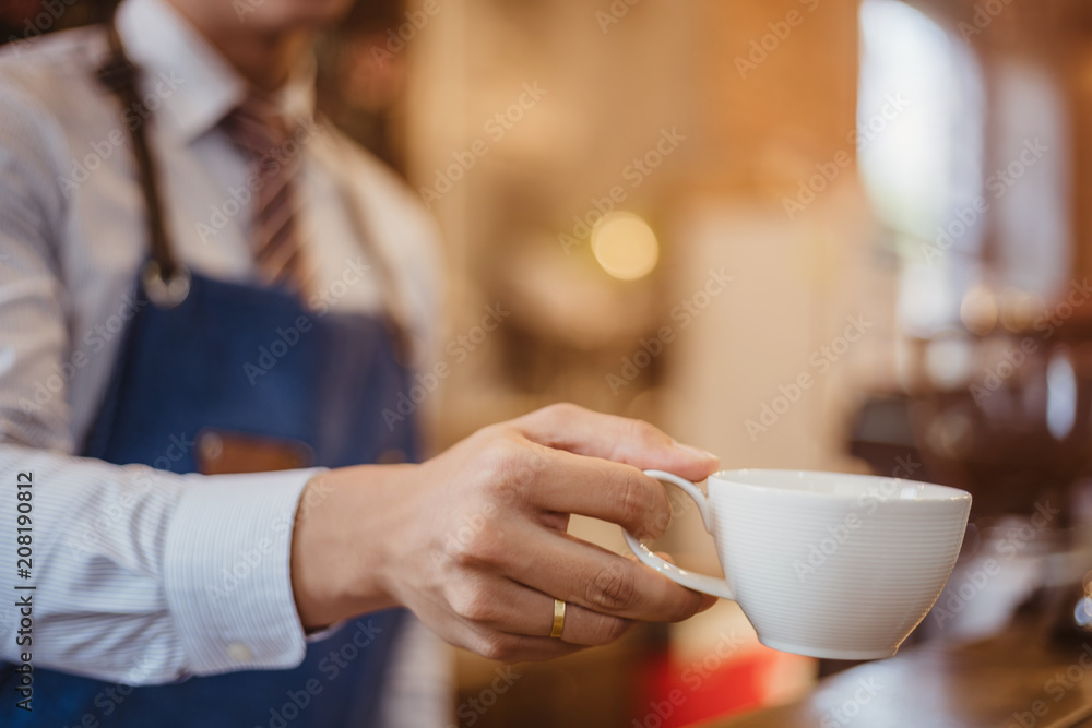 Special drink. Joyful nice handsome man wearing apron and holding a cup of tea while offering it to you Special drink. Joyful nice handsome man wearing apron and holding a cup of tea while offering it to you
