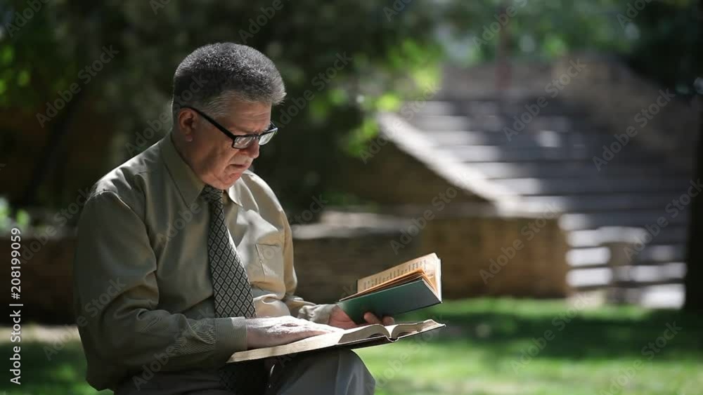 Profile of a smart scientist sitting on a bench and reading two books with a historicalstaircase behind his back in a city park in summer Profile of a smart scientist sitting on a bench and reading two books with a historicalstaircase behind his back in a city park in summer