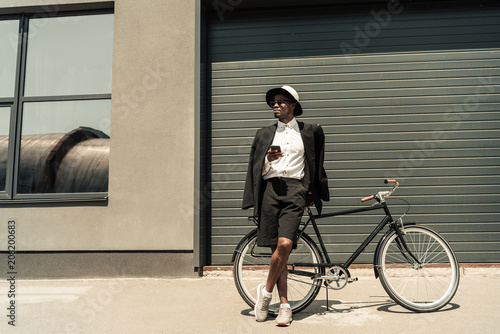 Stylish man wearing white shirt and jacket using smartphone while leaning on his bicycle
