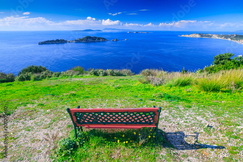 Awesome view from red wooden bench to the coastline sea bay with crystal clear azure water, distant islands in the calm warm sea. Beautiful summer seascape. Agios Stefanos cape. Afionas. Corfu. Greece