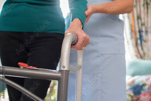 Senior Womans Hands On Walking Frame With Care Worker In Background