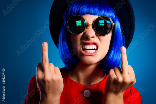 Closeup cropped portrait of nervous annoyed woman in wig eyewear showing teeth gesturing two fuck off signs with middle finger isolated on blue background