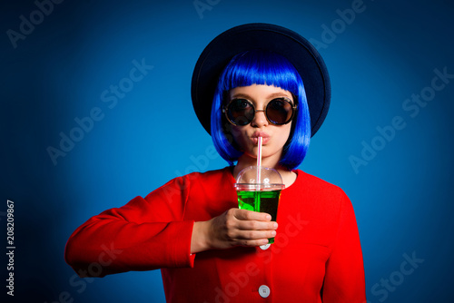 Portrait of pretty attractive girl in wig red outfit head wear eyewear having tasty sweet green cocktail in hand drinking from tubule with pout lips looking at camera isolated on blue background