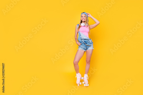 Portrait of slim pretty girl in denim overall baseball cap with headset on neck on roller skates holding hand in pocket of shorts looking at camera isolated on yellow background