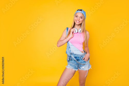 Portrait of pretty charming girl in cap with headset on neck holding hand in pocket of denim shorts looking at camera isolated on yellow background, pleasure delight enjoyment concept