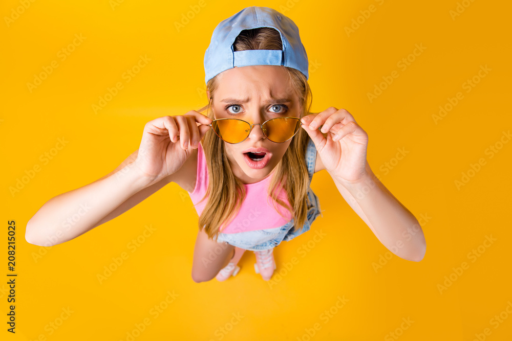 WTF! Top view portrait of sad disappointed girl looking out glasses on face holding eyelets with two hands isolated on yellow background WTF! Top view portrait of sad disappointed girl looking out glasses on face holding eyelets with two hands isolated on yellow background