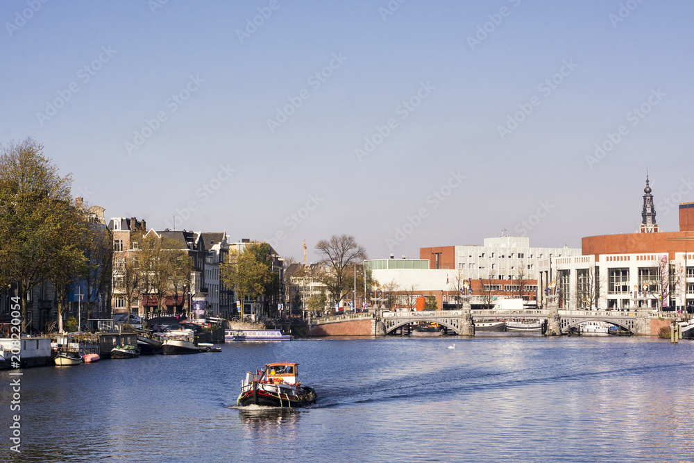 Obraz premium View on river Amstel in Amsterdam, capital of the Netherlands, with a boat, blue sky, trees and buildings