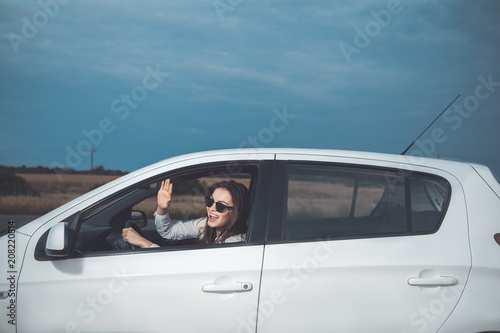Hello. Joyful young woman is driving a car and laughing. She is waving hand through the window. Picturesque meadow and sky on background 
