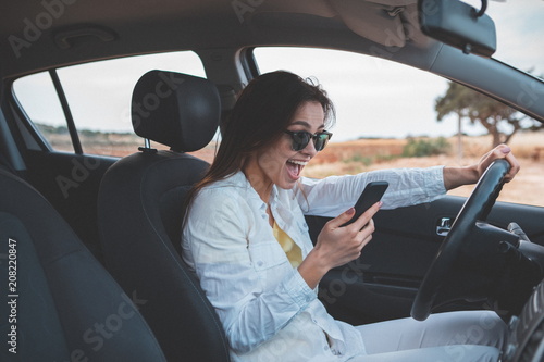 Excited young woman is reading message on smartphone and laughing. She is sitting at steering wheel of her car with relaxation. Beautiful natural landscape is on background  © Yakobchuk Olena