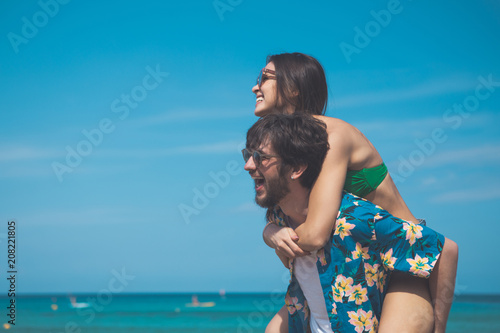 Side view of excited loving couple is enjoying summer vacation together. Man is holding woman on his back and smiling. They are looking at the sea with enjoyment  © Yakobchuk Olena