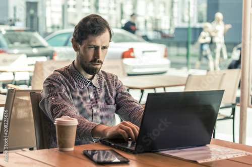 Young business man is using laptop sitting in cafe. A bearded focused guy works on a computer sitting at a table outdoors. 