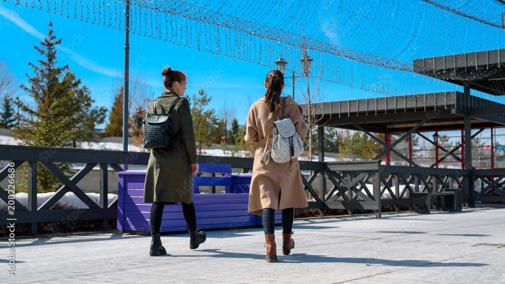 Two women in stylish spring coats and backpacks walk on the city embankment of Kazan. bottom rear view