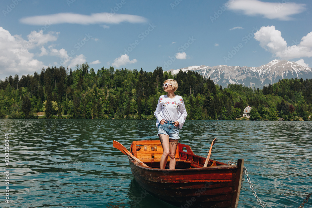beautiful young woman in sunglasses standing in boat at tranquil mountain lake, bled, slovenia