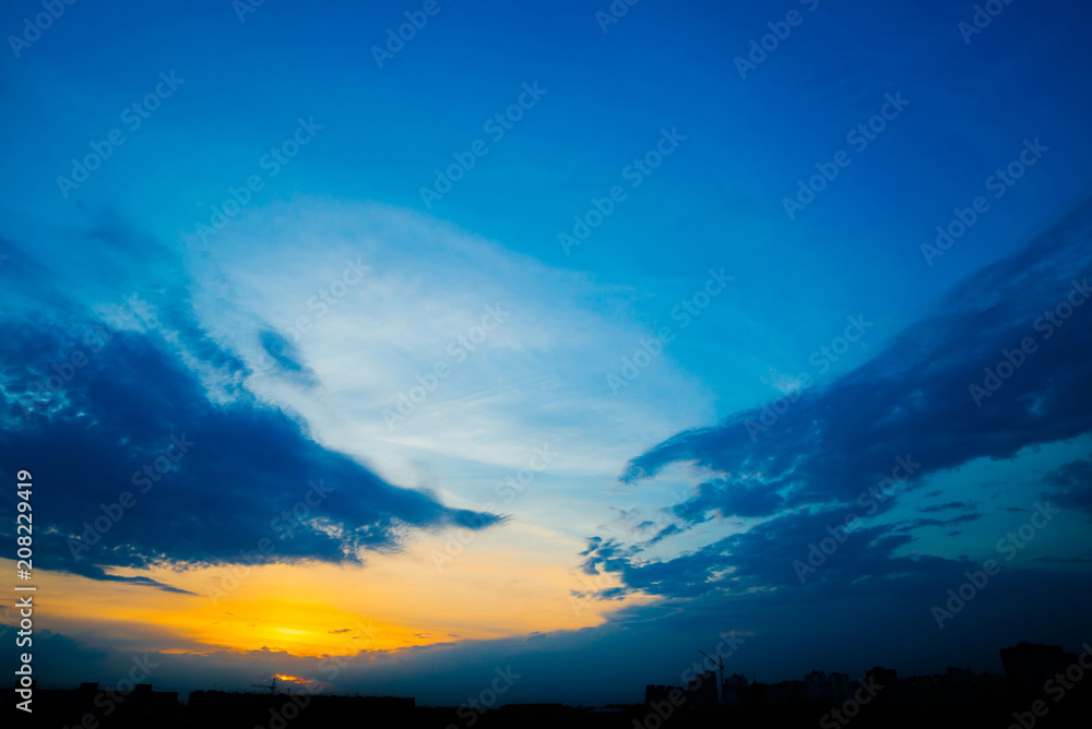 Atmospheric blue cloudy sky behind silhouettes of city buildings. Cobalt and orange background of sunrise with dense clouds and bright yellow sunny light for copy space. Cyan heaven above clouds. Atmospheric blue cloudy sky behind silhouettes of city buildings. Cobalt and orange background of sunrise with dense clouds and bright yellow sunny light for copy space. Cyan heaven above clouds.