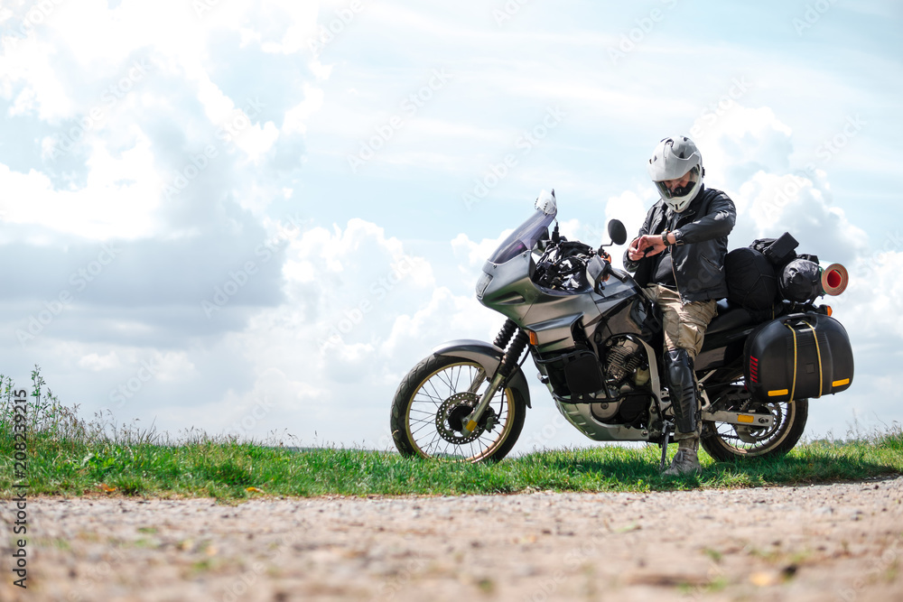 Rider Man looks at a watch and off road adventure motorcycles with side bags and equipment for long road trip, river and clouds on background, enduro travel touring concept