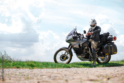 Rider Man looks at a watch and off road adventure motorcycles with side bags and equipment for long road trip, river and clouds on background, enduro travel touring concept © Sergey