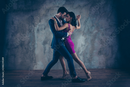 Full size body portrait of lovely attractive couple standing close in tango position face to face dancing, charming lady in purple tight dress gentlemen in tux isolated on grey background movement