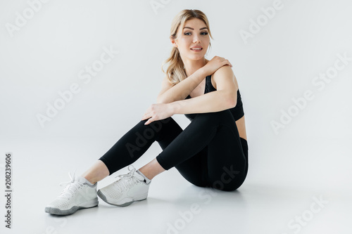smiling sportswoman in black sportswear sitting on floor isolated on grey