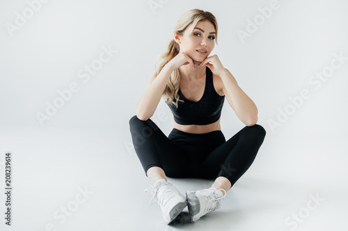attractive sportswoman in black sportswear sitting on floor isolated on grey