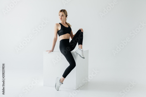 sportive woman in black sportswear sitting on white cube on grey background