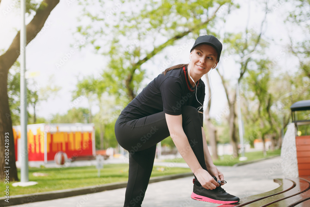 Young athletic beautiful brunette girl in black uniform and cap with headphones listening music, tying shoelaces before running, training on bench in city park outdoors. Fitness, healthy lifestyle.