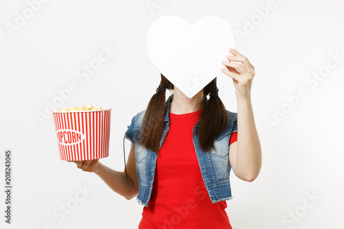 Portrait of young brunette woman in casual clothes watching movie film, holding bucket of popcorn and covering face with white heart with copy space isolated on white background. Emotions in cinema. photo