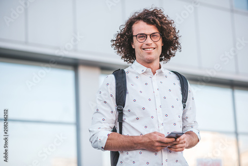 Handsome young happy Caucasian male wearing trendy spectacles and shirt looking at the camera with his smart phone and backpack, posing outdoor. People, lifestye and modern technology communication. photo