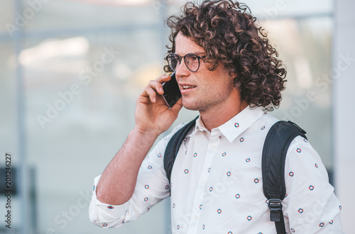 Close up portrait of stylish curly hair male in trendy spectacles standing outdoors while holding backpack and talking by his smart phone outdoor on the city street. People, technology, lifestyle. photo