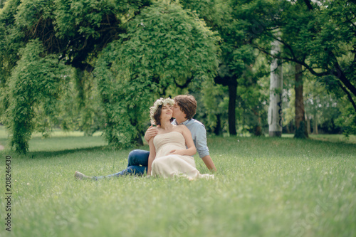 Fototapeta Naklejka Na Ścianę i Meble -  Happy pregnant woman and her husband sitting on the blanket in the green meadow in the park. Sunny summer day