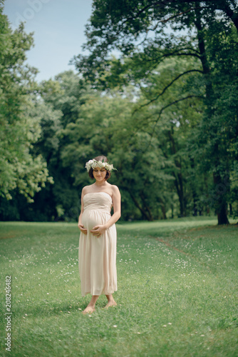 Portarit of tender pregnant woman in beautiful creme dress and flower wreath walking on the reen grass in the sunny summer park