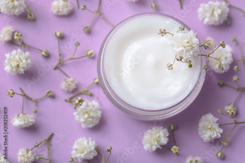 Jar of body cream and beautiful flowers on color background
