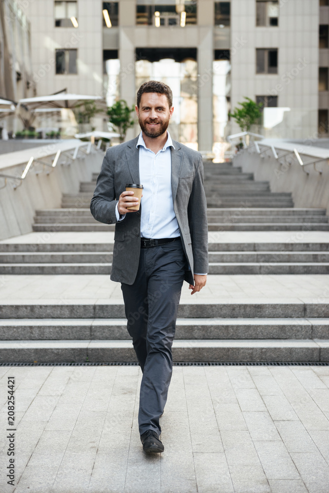 Photo of pleased entrepreneur man 40s in gray suit walking down stairs of modern business center, with takeaway coffee in hand Photo of pleased entrepreneur man 40s in gray suit walking down stairs of modern business center, with takeaway coffee in hand