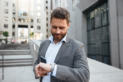 Photo of adult handsome man in gray suit holding mobile phone, and looking at wrist watch while standing in front of modern office building