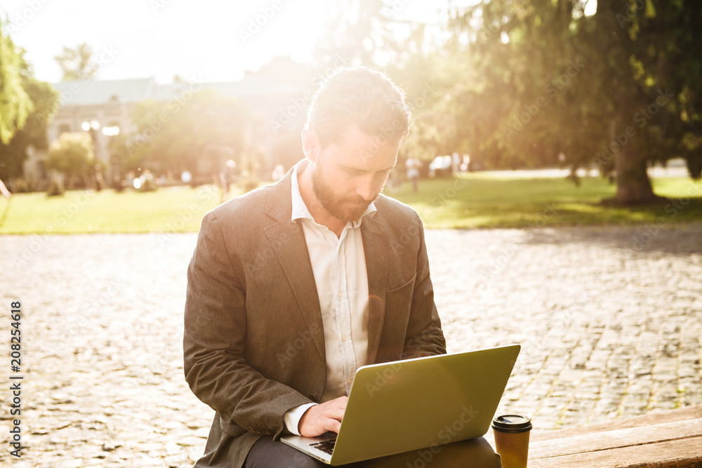 Image of serious businessman in gray classical suit, sitting in park with takeaway coffee and working on silver laptop on sunny day