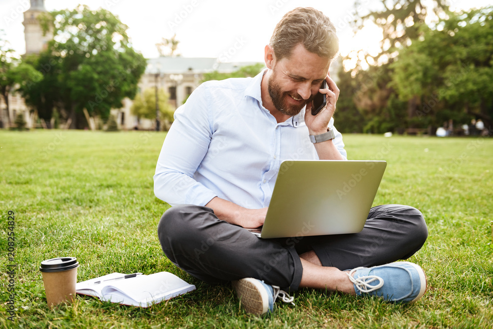 Photo of adult european man in business clothing, sitting on grass in park with legs crossed and having business call while working on silver laptop