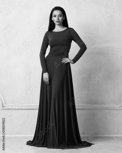 Young woman with long brunette hair dressed in elegant evening blue maxi dress with long sleeves posing in studio. Gorgeous female model standing against white wall decorated with classic moldings.