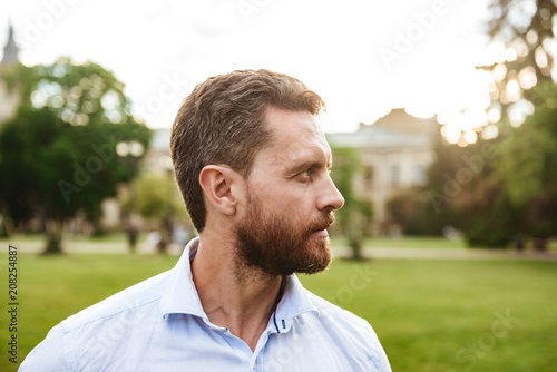 Portrait closeup of serious man 40s in white shirt, looking aside during walk in green park while standing over old building background