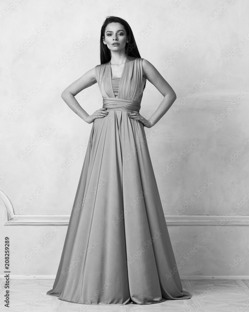 Fototapeta premium Beautiful long haired young woman dressed in stylish purple bandeau maxi dress posing against white wall on background. Elegant brunette female model demonstrating evening outfit in studio.