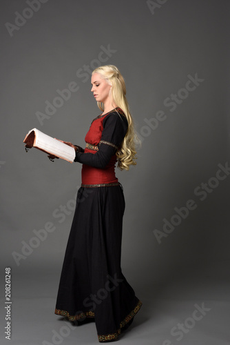  full length portrait of pretty blonde lady wearing a red and black fantasy medieval gown, holding a book. standing pose on grey background.