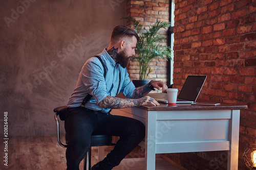 Handsome tattooed hipster in a shirt and suspenders sitting at the desk, working on a laptop, holds takeaway coffee in an office with a loft interior.