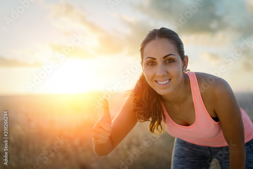 Successful female runner taking a rest during outdoor running workout at sunset. Female athlete doing success thumbs up geture. Sport and exercise motivation concept.